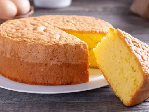 Un moelleux gâteau de Savoie, parfait pour la conservation, avec une part coupée. Des œufs et un fouet en arrière-plan suggèrent sa préparation.