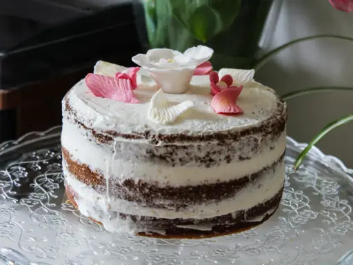 Un délicieux layer cake fleuri, avec des pétales de roses en sucre roses et blancs, posé sur un plat en verre orné.