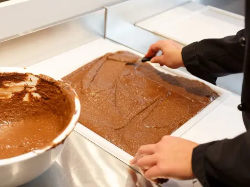 Un chef étale une pâte chocolatée onctueuse dans un moule. C'est la base du praliné, une gourmandise qui fait rêver.