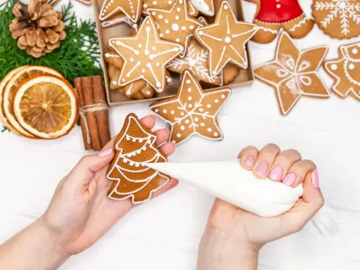 Mains décorant un biscuit sapin avec du glaçage royal. Autour, des étoiles et des décorations de Noël.