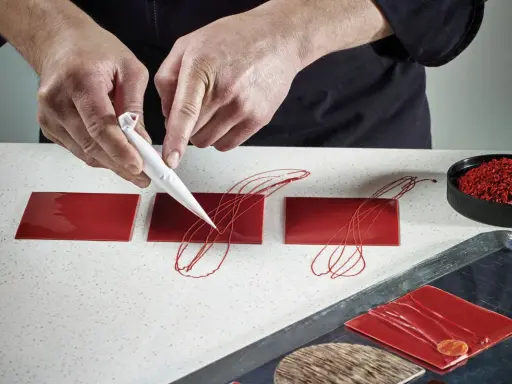 Pâtissier décorant des carrés de chocolat rouge avec un cornet. Le papier guitare, c'est quoi ? C'est ce support qui permet ces décorations fines.