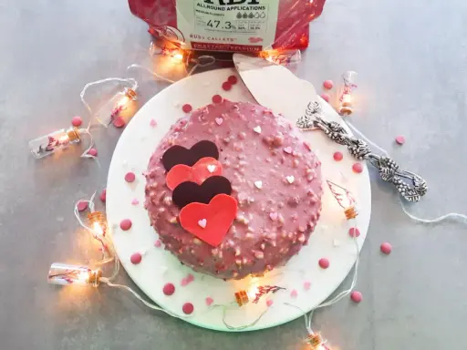 Gâteau rose orné de cœurs, enrobé de chocolat ruby croquant. Guirlande lumineuse et confettis ajoutent une touche festive.