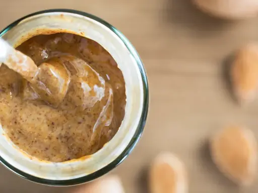 Une cuillère plonge dans un pot de purée d'amande maison, crémeuse et onctueuse. Des amandes sont disposées autour.
