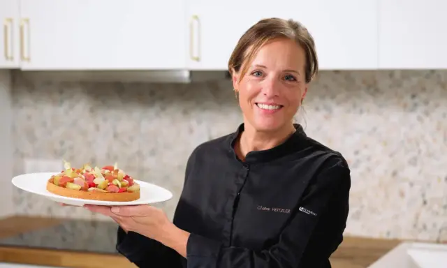 Pâtissière souriante tenant une tartelette aux fruits, peut-être préparée avec de la pectine ou de la gélatine pour une texture parfaite.