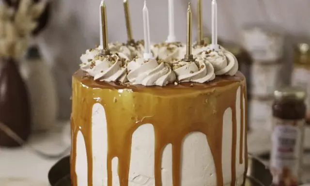 Gâteau d'anniversaire à étages, nappé de caramel beurre salé, décoré de crème et de noisettes concassées, avec des bougies dorées et blanches.
