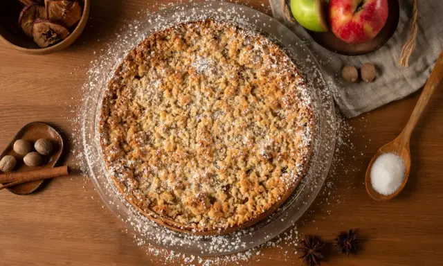 Un délicieux crumble pomme, saupoudré de sucre glace, entouré de pommes fraîches, de noix de muscade et de bâtons de cannelle.