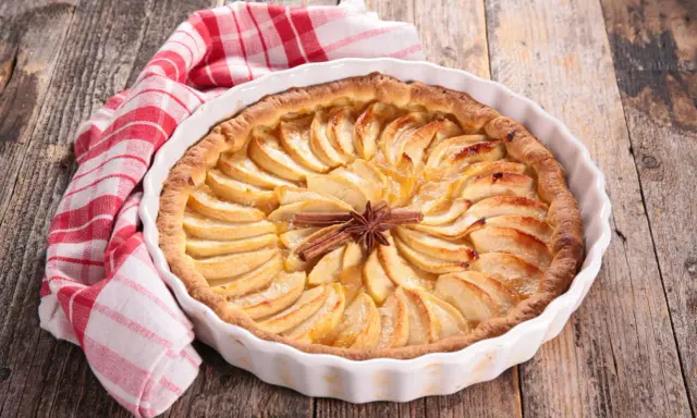 Tarte aux pommes dorée, décorée de bâtons de cannelle et d'anis étoilé. Une serviette à carreaux rouges et blancs est posée à côté. Idéale pour la conservation.