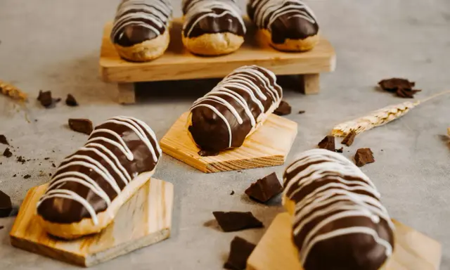 Éclairs gourmands avec un glaçage éclair chocolat brillant, décorés de fines rayures blanches. Quelques morceaux de chocolat et une tige de blé complètent la scène.