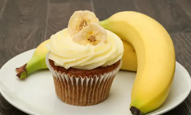 Un délicieux cupcake banane, orné de fines tranches de banane séchée, repose sur une assiette blanche, à côté de bananes fraîches.