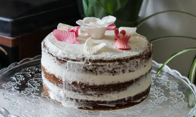Un délicieux layer cake fleuri, avec des pétales de roses en sucre roses et blancs, posé sur un plat en verre orné.