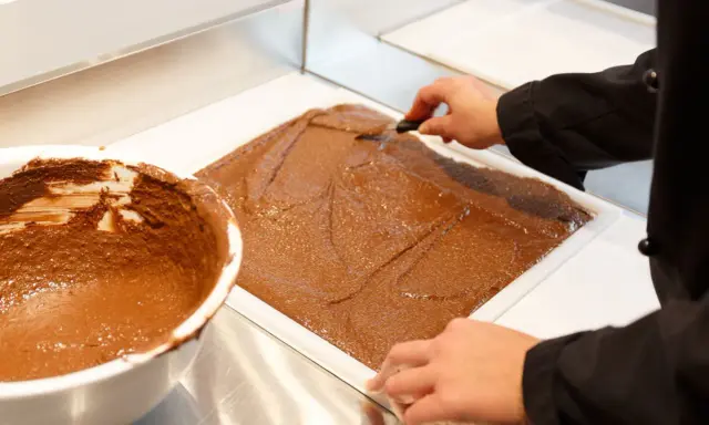 Un chef étale une pâte chocolatée onctueuse dans un moule. C'est la base du praliné, une gourmandise qui fait rêver.