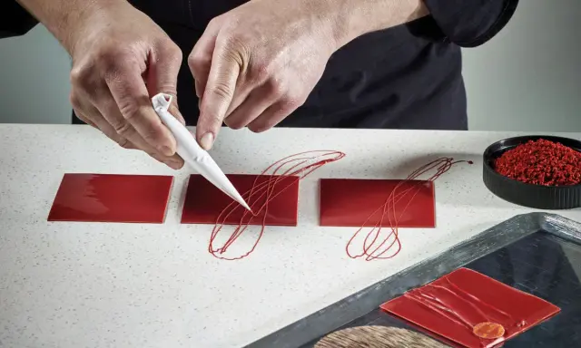 Pâtissier décorant des carrés de chocolat rouge avec un cornet. Le papier guitare, c'est quoi ? C'est ce support qui permet ces décorations fines.