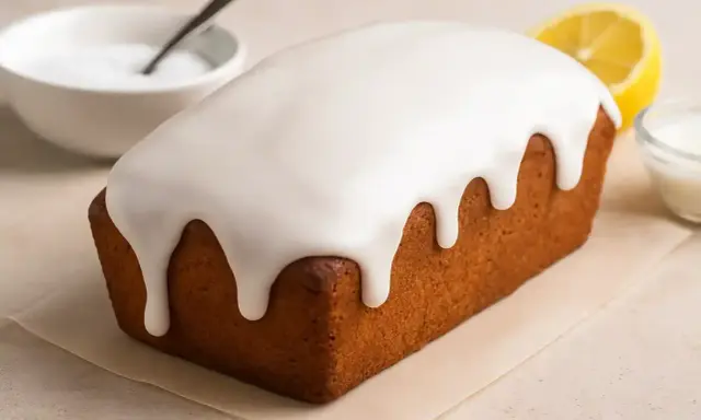Un délicieux cake au citron recouvert d'un glaçage sucre glace facile, avec une tranche de citron et un bol d'ingrédients à proximité.