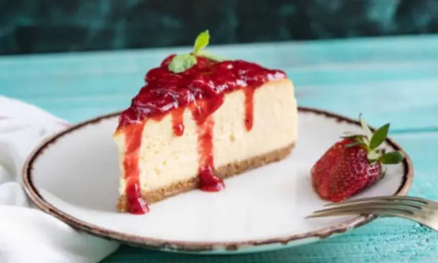 Tranche de cheesecake à la ricotta sans cuisson, nappée de coulis de fraises et décorée d'une feuille de menthe. Une fraise entière et une fourchette complètent la présentation.