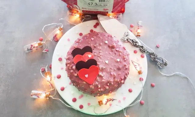 Gâteau rose orné de cœurs, enrobé de chocolat ruby croquant. Guirlande lumineuse et confettis ajoutent une touche festive.