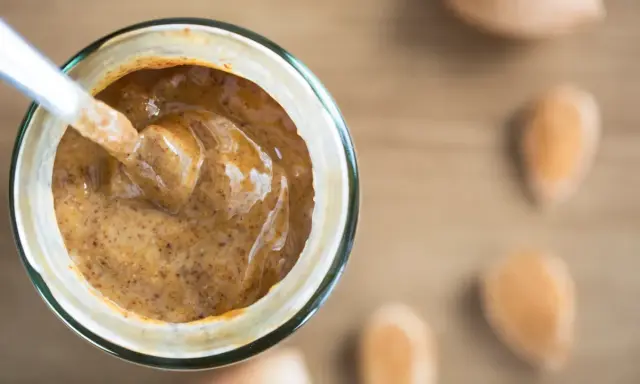 Une cuillère plonge dans un pot de purée d'amande maison, crémeuse et onctueuse. Des amandes sont disposées autour.