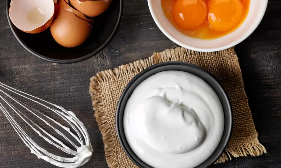 Préparation d'une meringue : blancs en neige, jaunes d'œufs, coquilles et fouet sur fond de bois sombre.