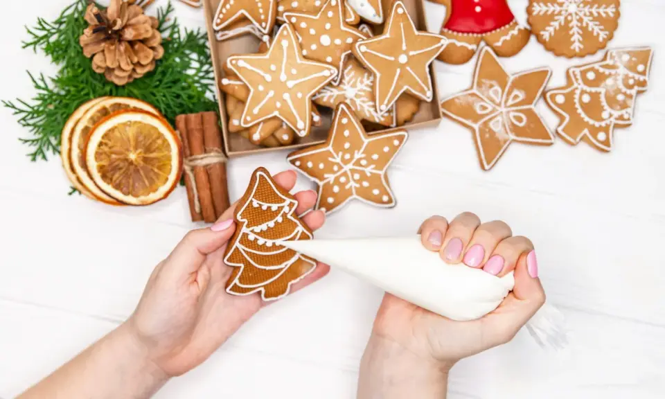 Mains décorant un biscuit sapin avec du glaçage royal. Autour, des étoiles et des décorations de Noël.