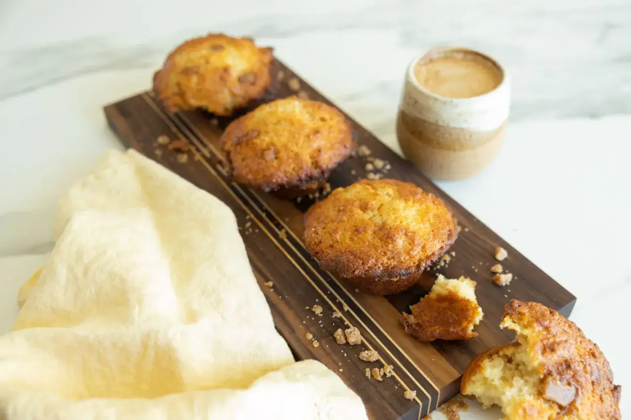 Trois muffins banane caramel dor&eacute;s sur une planche en bois, avec une tasse de caf&eacute; et un tissu jaune.