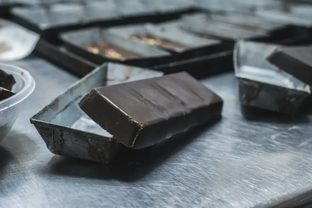 Une barre de chocolat sombre repose sur une petite boîte métallique. Des copeaux de chocolat sont visibles dans un bol à gauche.