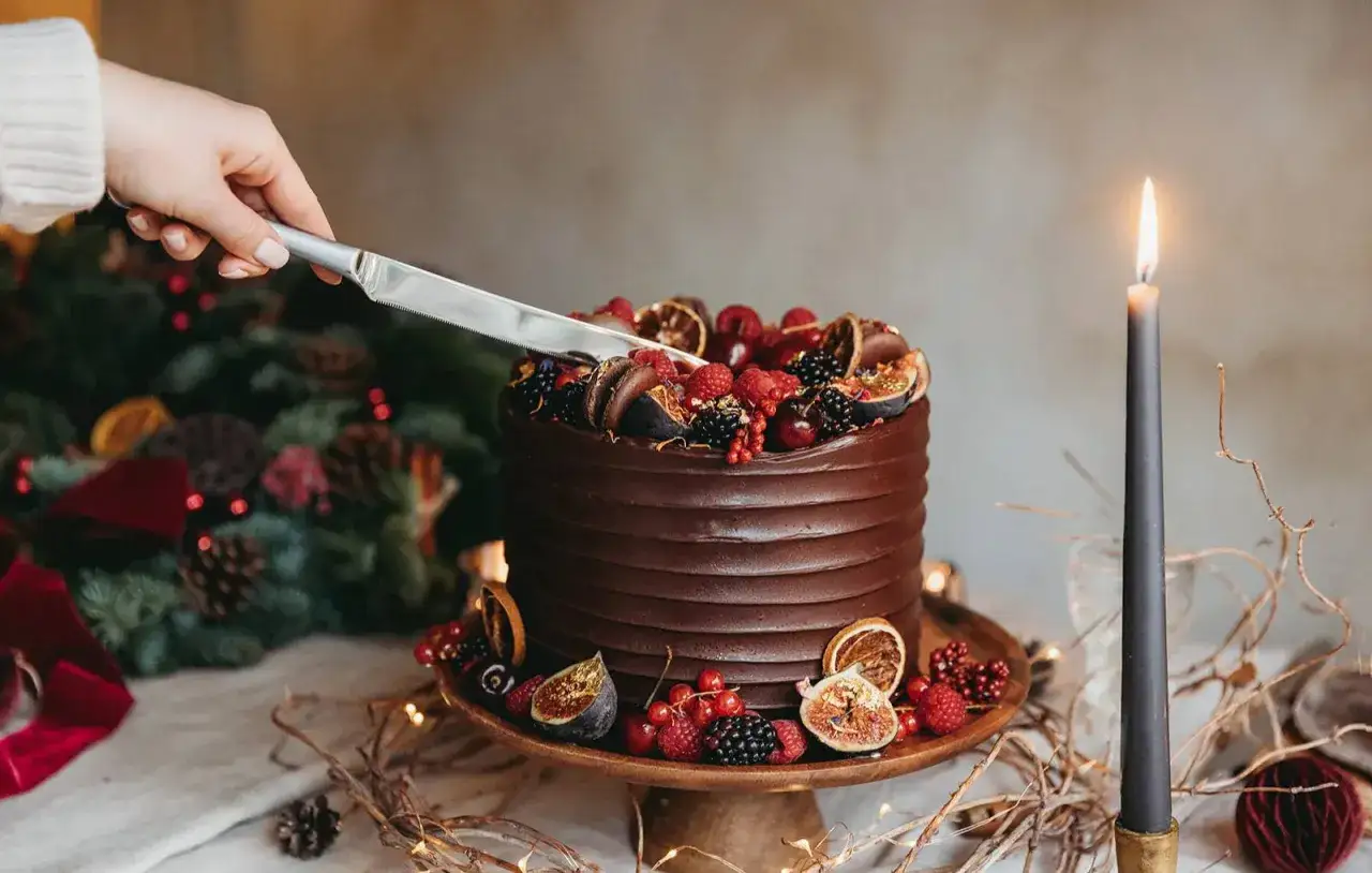 Une main coupe un magnifique gateau d'anniversaire chocolat, décoré de fruits frais et de figues. Une bougie allumée complète la scène festive.