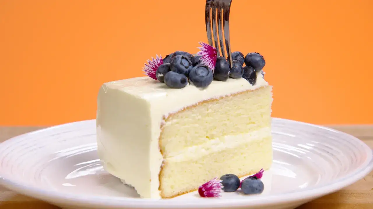 Tranche de gâteau moelleux, recouverte d'une onctueuse butter cream et garnie de myrtilles fraîches. Une fourchette s'apprête à déguster ce délice.