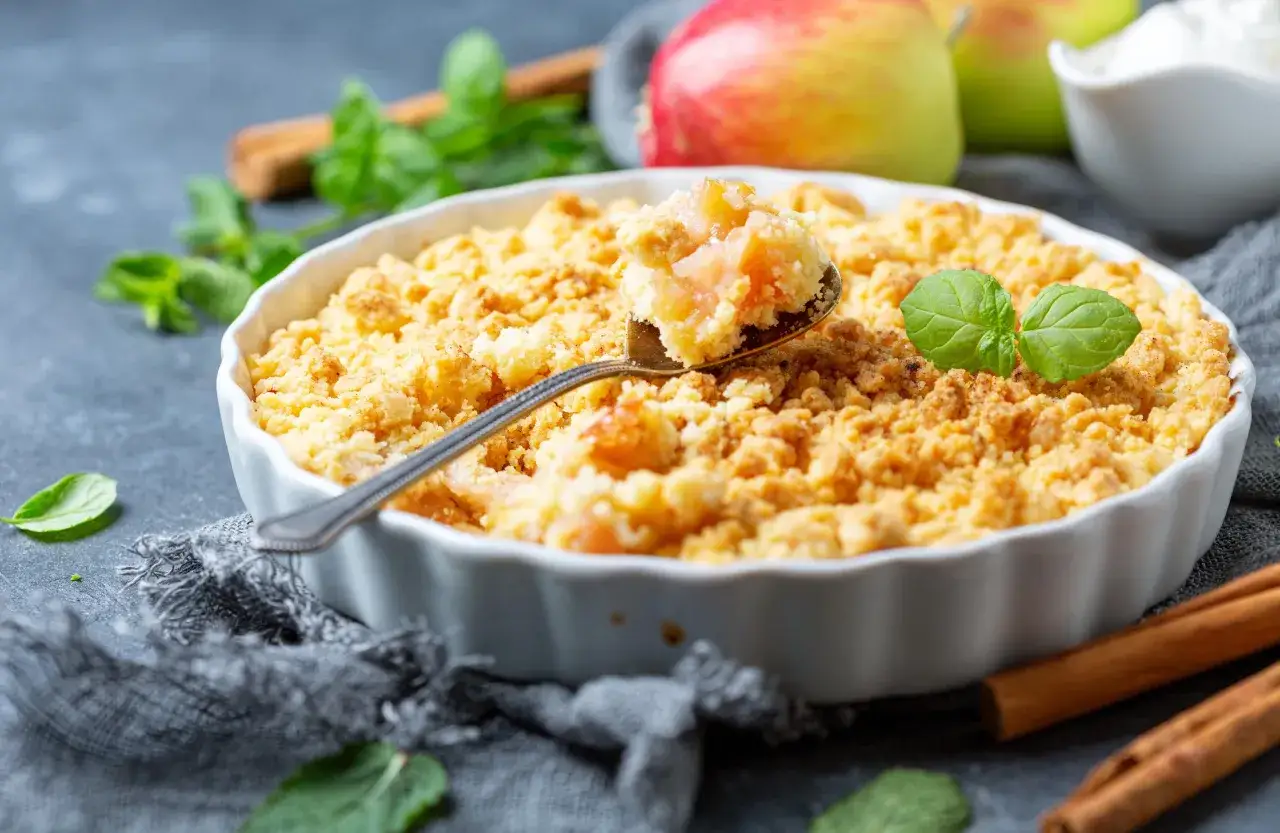 Un d&eacute;licieux crumble pomme dor&eacute;, garni de feuilles de menthe, avec des pommes et des b&acirc;tons de cannelle en arri&egrave;re-plan.