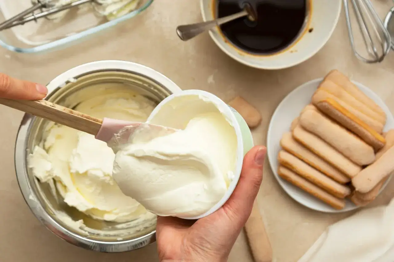 Préparation d'une recette mascarpone : on ajoute le fromage crémeux dans un bol avec une spatule. Des biscuits à la cuillère et du café sont prêts.