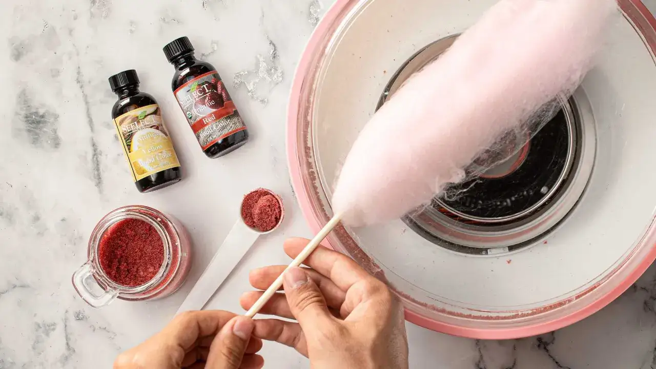 Comment faire du sucre à barbe à papa : des colorants alimentaires rouge et jaune, du sucre en poudre et une machine à barbe à papa en action.