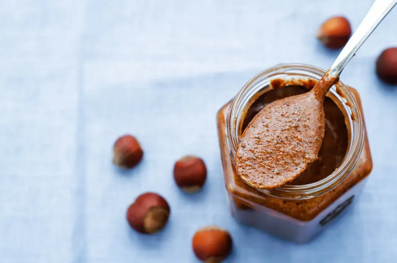 Une cuillère de praliné noisette sort d'un pot, entouré de noisettes. Le praliné c'est quoi ? Une pâte onctueuse et gourmande.