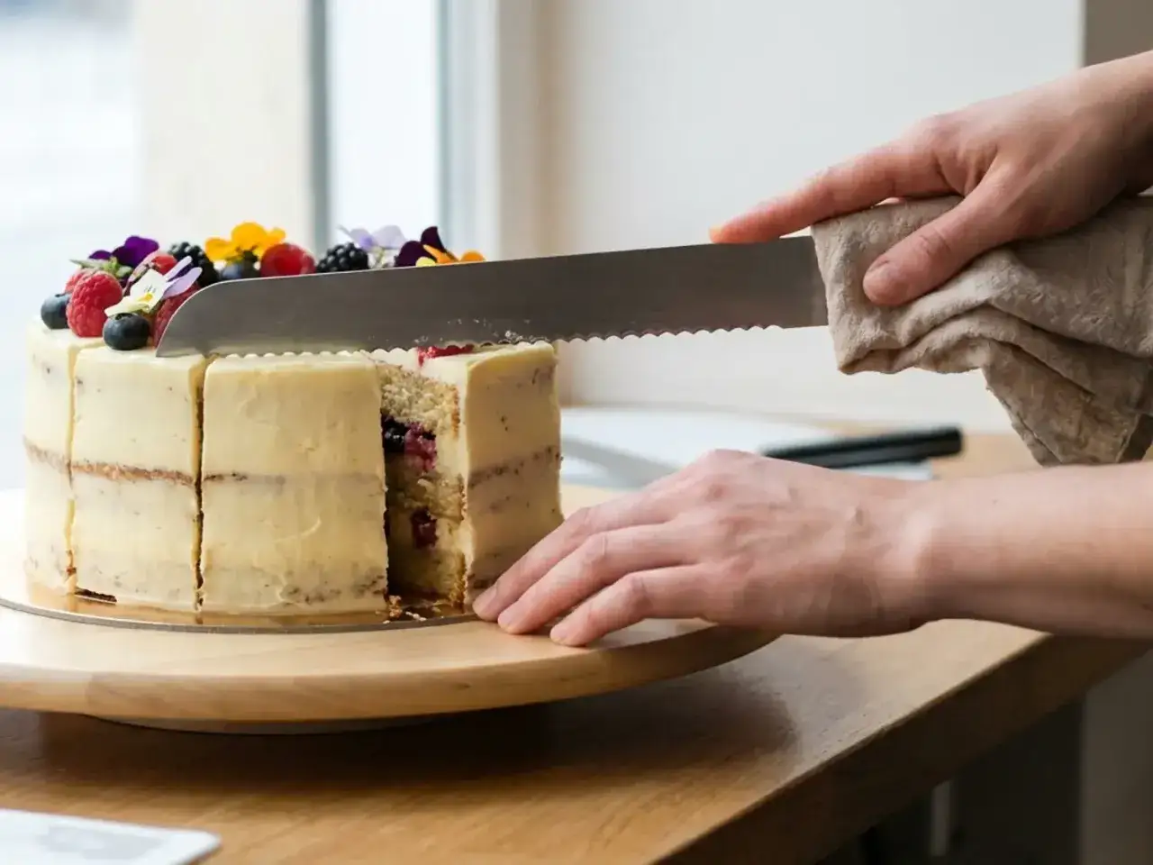 Comment couper un layer cake : une main tient un torchon pour essuyer le couteau denté avant de trancher ce beau gâteau garni de fruits rouges et de fleurs.
