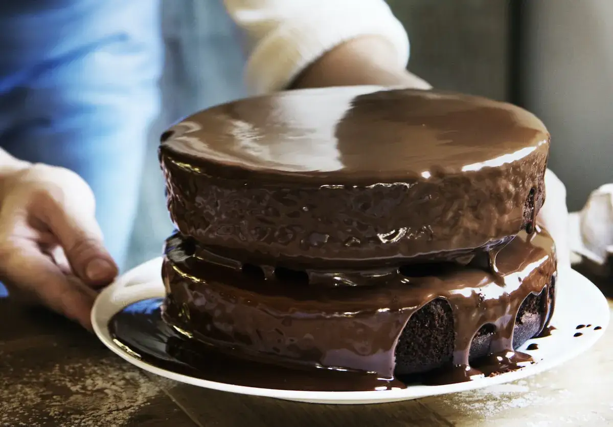 Un gâteau au chocolat à deux étages, nappé d'un glaçage brillant. Une main le présente, prêt à être dégusté, un vrai délice pour la conservation des gourmands.