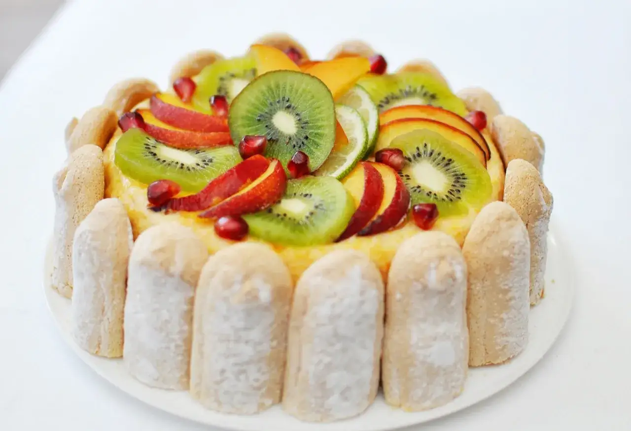 Charlotte aux fruits colorée, garnie de kiwis, pêches, grenades et quartiers de citron vert, entourée de biscuits à la cuillère saupoudrés de sucre glace.