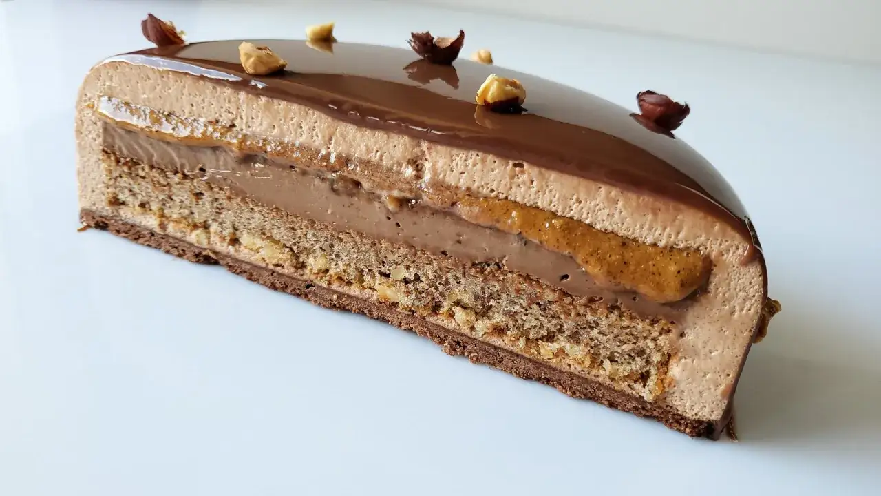 Tranche d'un tiramisu noisette aux couches de mousse chocolat, caramel et biscuit croustillant, décoré de noisettes entières.
