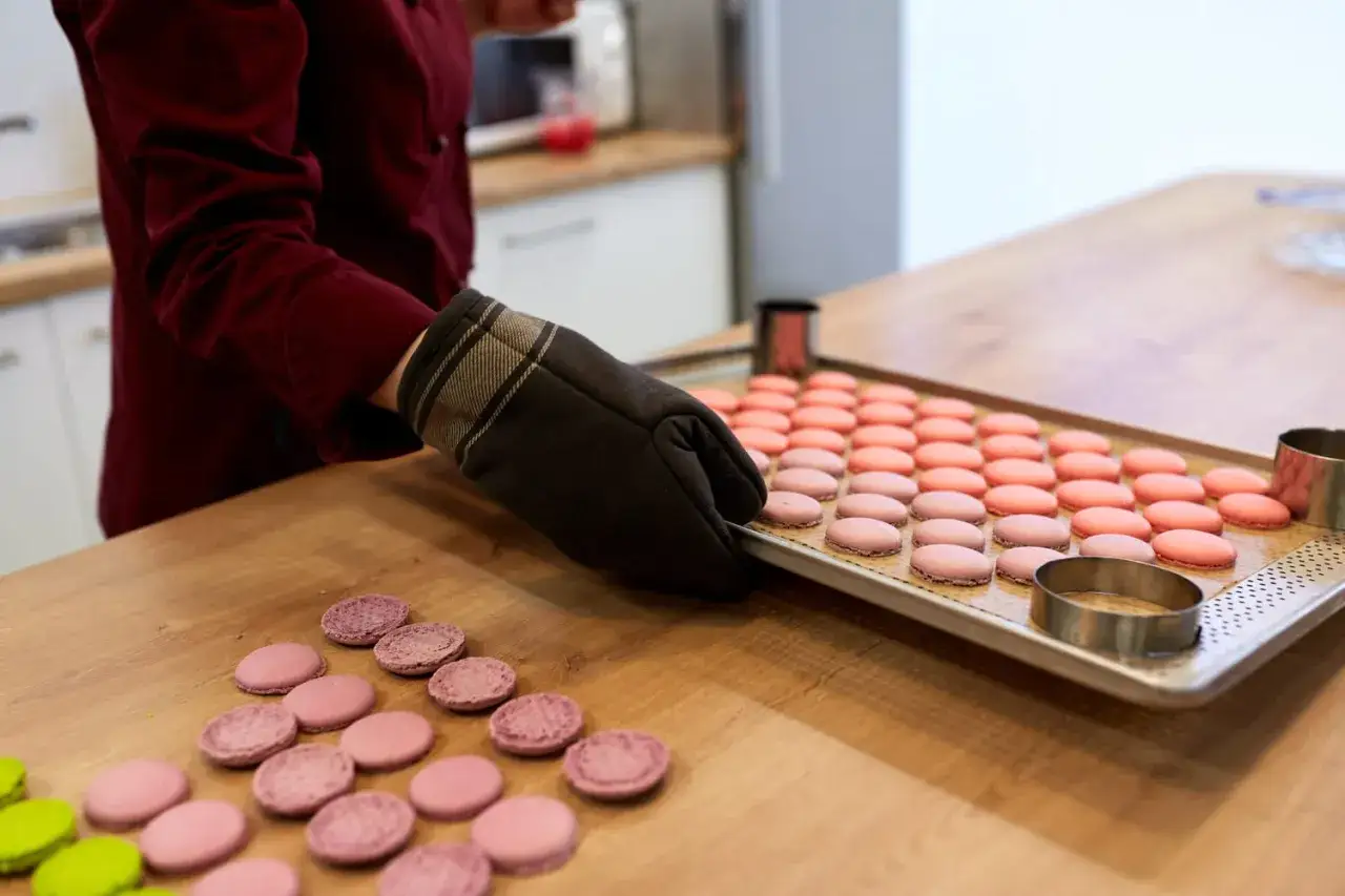 Préparation pour la cuisson macaron four traditionnel. Une main gantée dépose une plaque de macarons roses sur un plan de travail en bois.