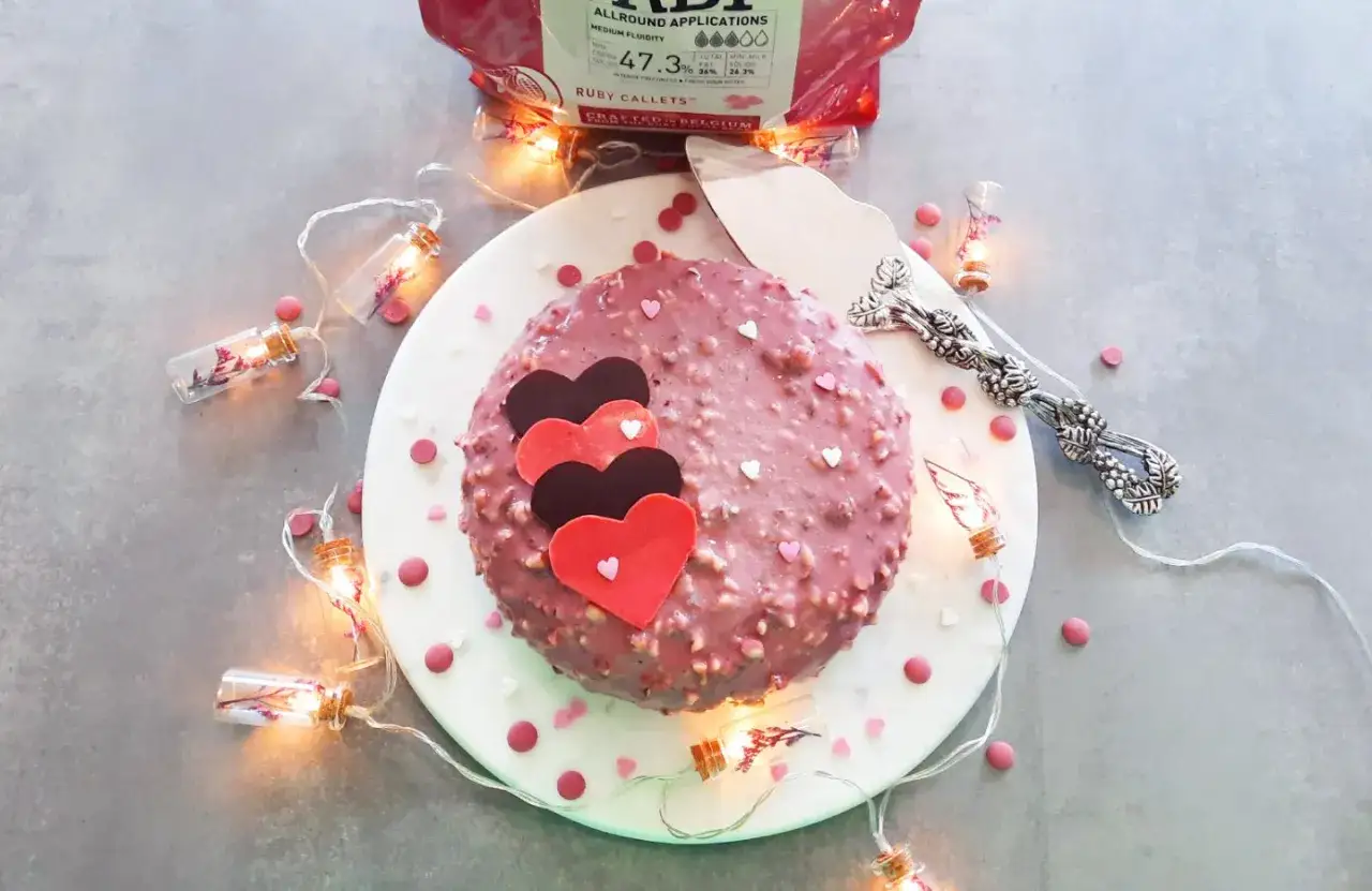 Gâteau rose orné de cœurs, enrobé de chocolat ruby et de noix. Guirlande lumineuse et confettis en forme de cœur l'entourent.