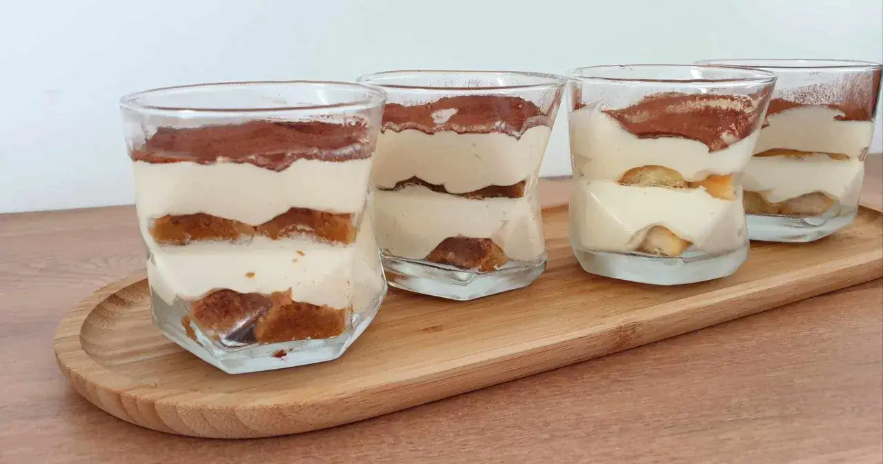 Quatre verrines de tiramisu chantilly mascarpone, saupoudr&eacute;es de cacao, sur un plateau en bois.