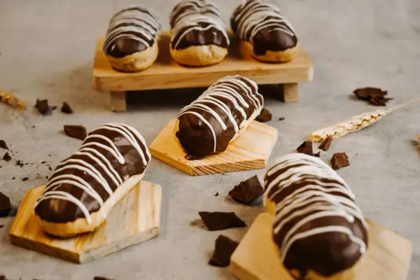 Éclairs gourmands avec un glaçage éclair chocolat brillant, décorés de fines rayures blanches. Quelques morceaux de chocolat et une tige de blé complètent la scène.