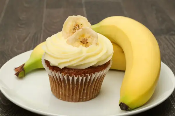 Un délicieux cupcake banane, orné de fines tranches de banane séchée, repose sur une assiette blanche, à côté de bananes fraîches.