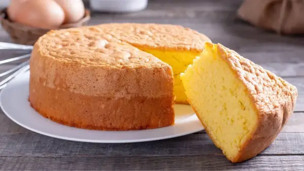 Un moelleux gâteau de Savoie, parfait pour la conservation, avec une part coupée. Des œufs et un fouet en arrière-plan suggèrent sa préparation.