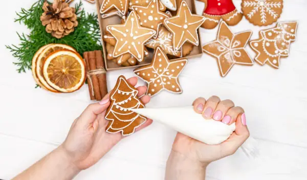 Mains décorant un biscuit sapin avec du glaçage royal. Autour, des étoiles et des décorations de Noël.