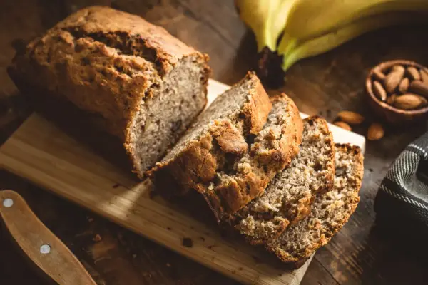 Tranches de banana bread moelleux sur une planche en bois, avec des bananes et des amandes en arrière-plan.