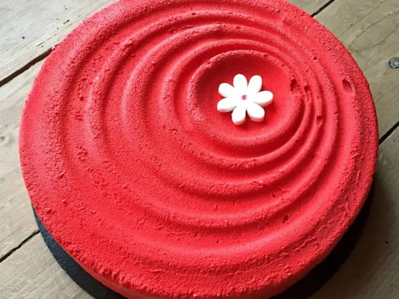 Un tiramisu fraise rhubarbe d'un rouge éclatant, décoré d'une petite fleur blanche en son centre, sur un fond de bois.